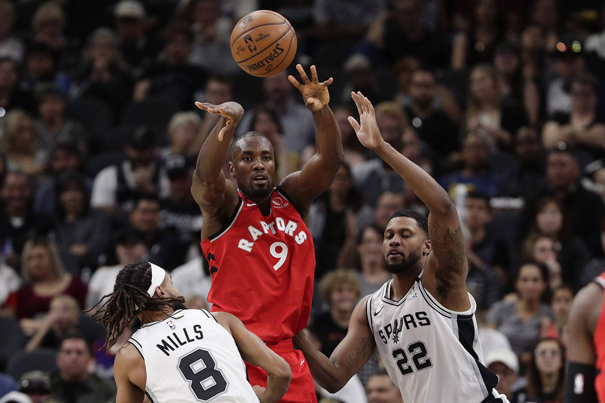 FILE – Toronto Raptors center Serge Ibaka (9) passes the ball over San Antonio Spurs guard Patty Mills (8) and forward Rudy Gay (22) during the second half of an NBA basketball game in San Antonio, Sunday, Jan. 26, 2020. (AP Photo/Eric Gay)