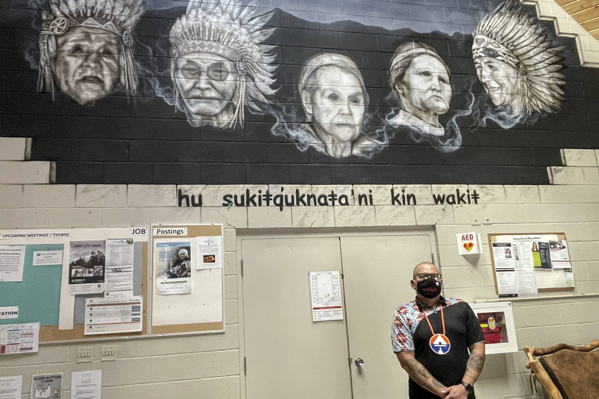 Nasukin Jason Louie of the Lower Kootenay Band poses under the mural in the administration building. The mural depicts past elders David Luke, Wilfred Jacobs, Isobel Louie, Charlotte Basil, and Louis White. (Photo by Kelsey Yates)