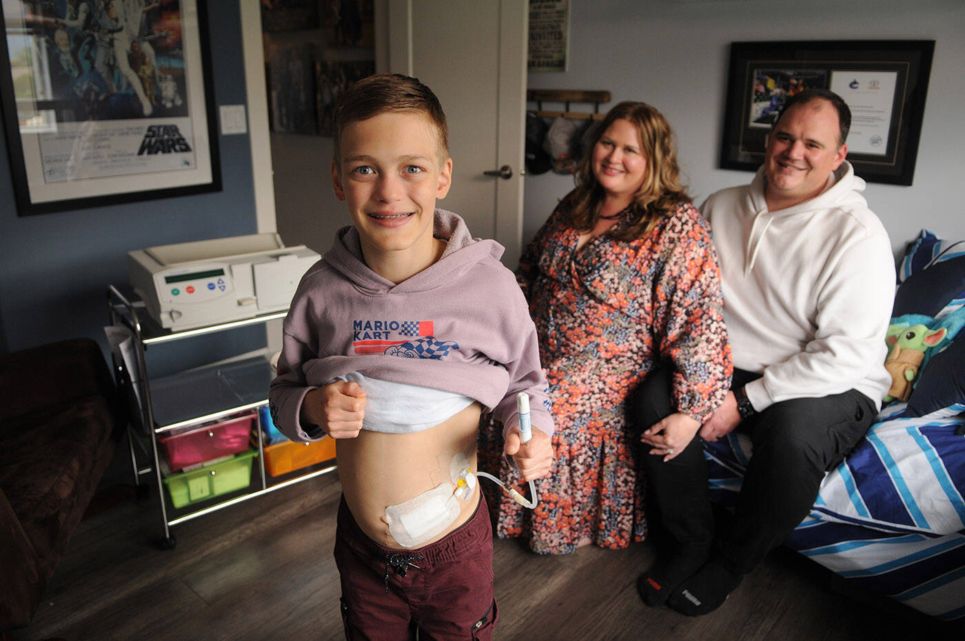 Thirteen-year-old Madden Wicker-Vriend, seen here with his parents Eryn and Jesse on May 27, is on dialysis at home in Chilliwack and is looking for a living kidney donor. He’s holding his catheter and behind him is his dialysis machine. (Jenna Hauck/ Chilliwack Progress)